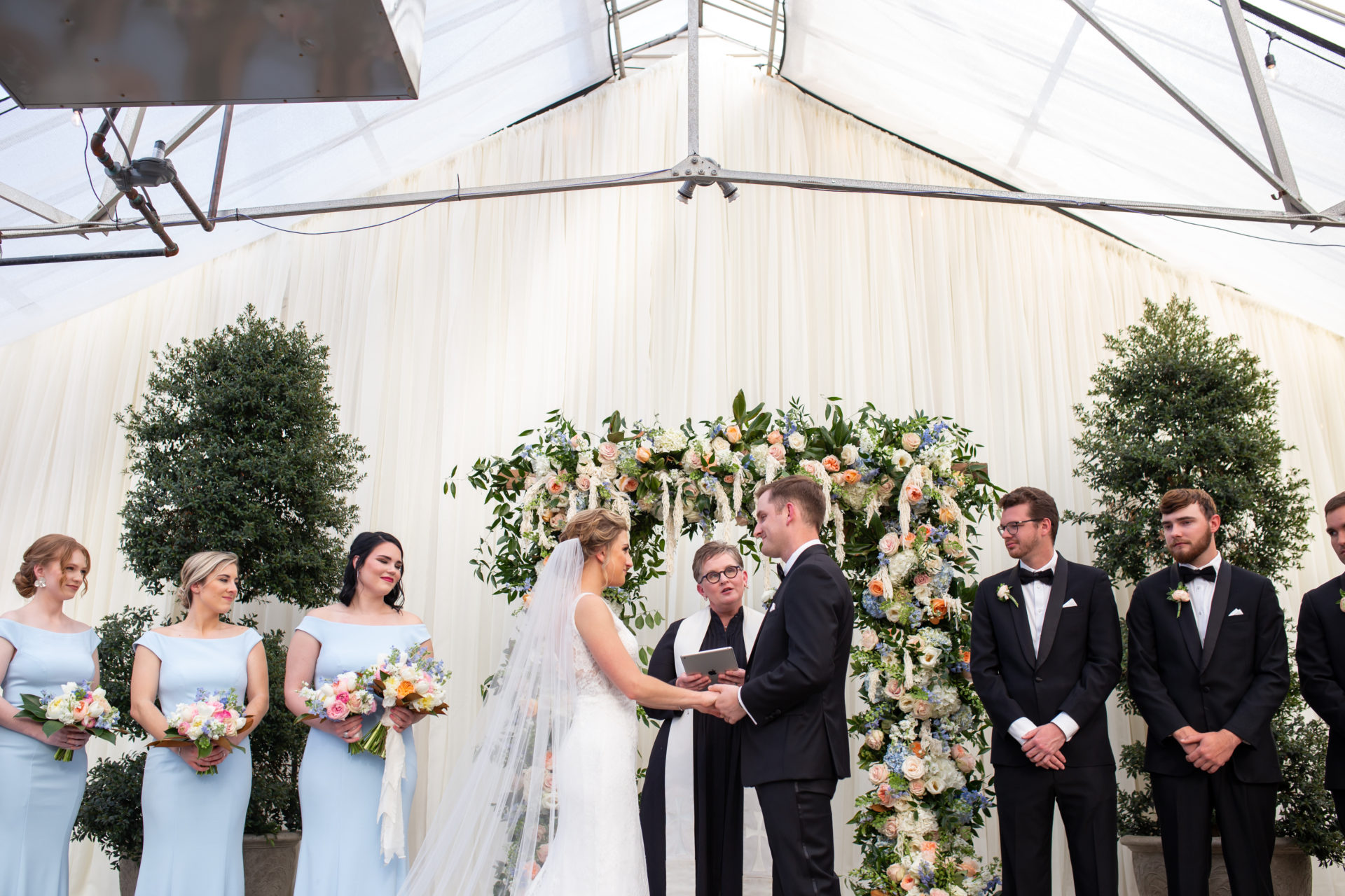 blonde bride and groom in black tux marrying under bright floral arch